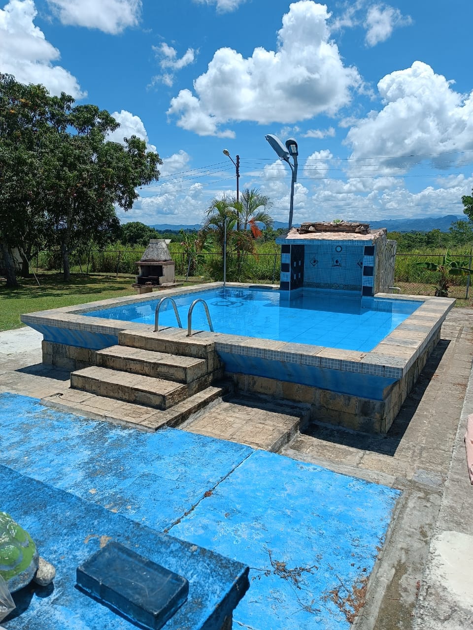 A private swimming pool is surrounded by a blue-tiled deck, featuring steps leading down into the water. Lush greenery and distant hills are visible under a bright sky dotted with clouds, creating an inviting outdoor environment.