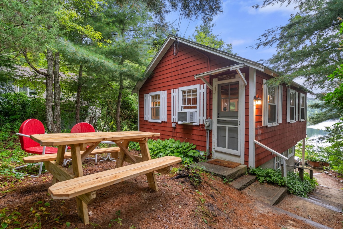 A quaint red cottage is nestled among trees, with a front porch accessed by stone steps. A picnic table sits on a natural ground, accompanied by two bright red Adirondack chairs, providing a space for outdoor relaxation and dining near the lake.