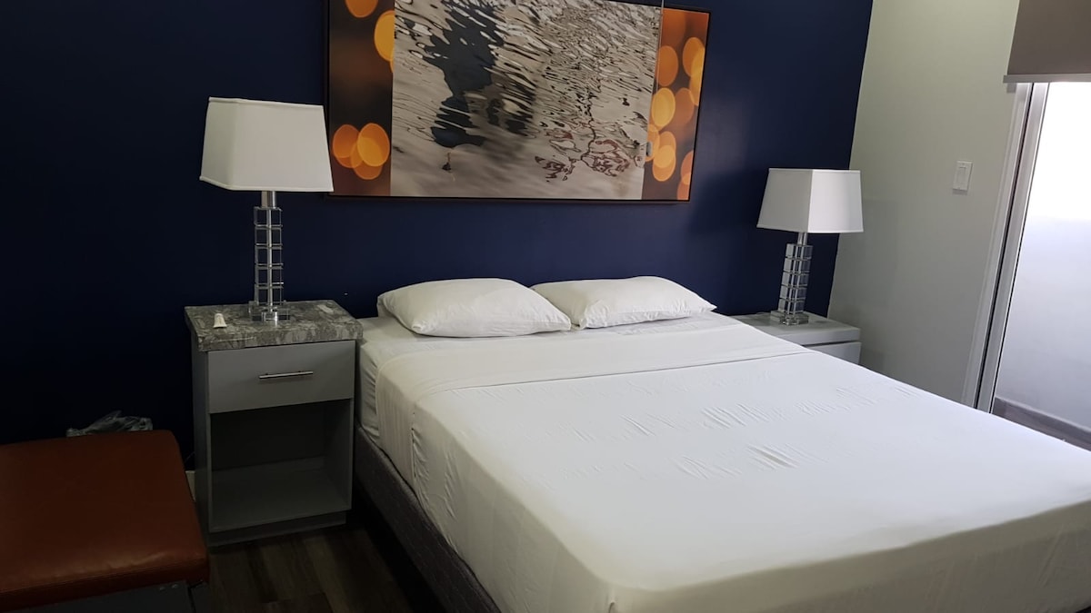 A well-appointed sleeping area features a spacious bed with a neatly arranged white bedspread. Two bedside tables with modern lamps flank the bed, complementing the blue accent wall and a decorative artwork above. Natural light enters from a nearby window.
