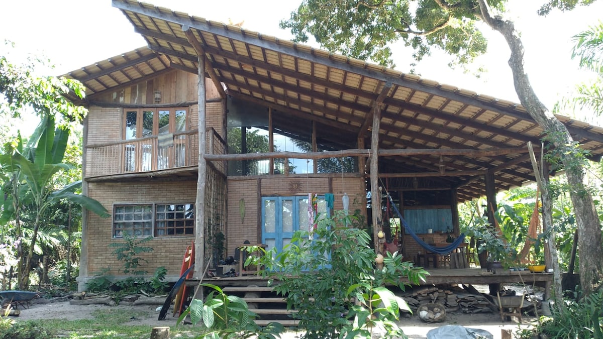 A rustic house constructed from wood and eco-friendly bricks is surrounded by lush greenery. Large windows allow ample natural light into the living spaces. A spacious deck offers room for relaxation, while a hammock is visible, inviting tranquil moments outdoors.