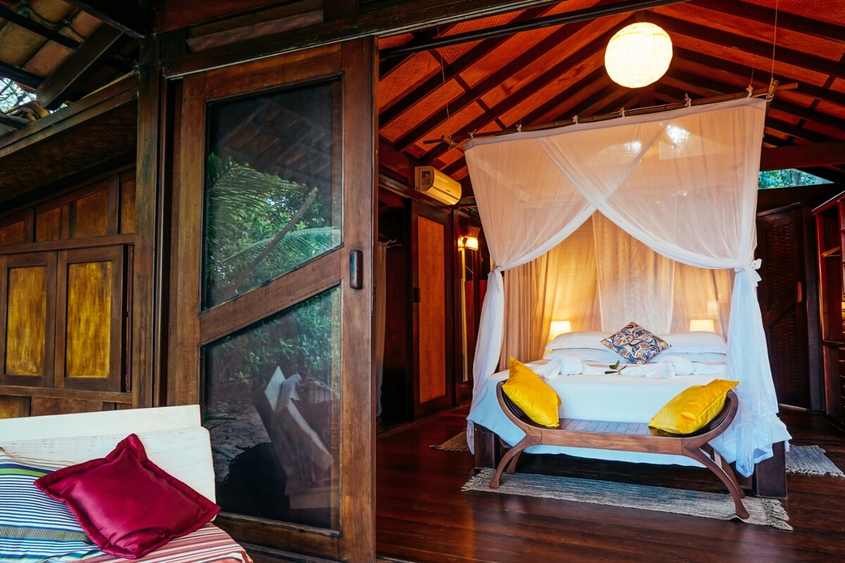 A serene sleeping area features a spacious bed draped with a sheer canopy, inviting soft lighting from bedside lamps. Natural wood finishes enhance the rustic charm, framed by large glass doors that open to the surrounding greenery.