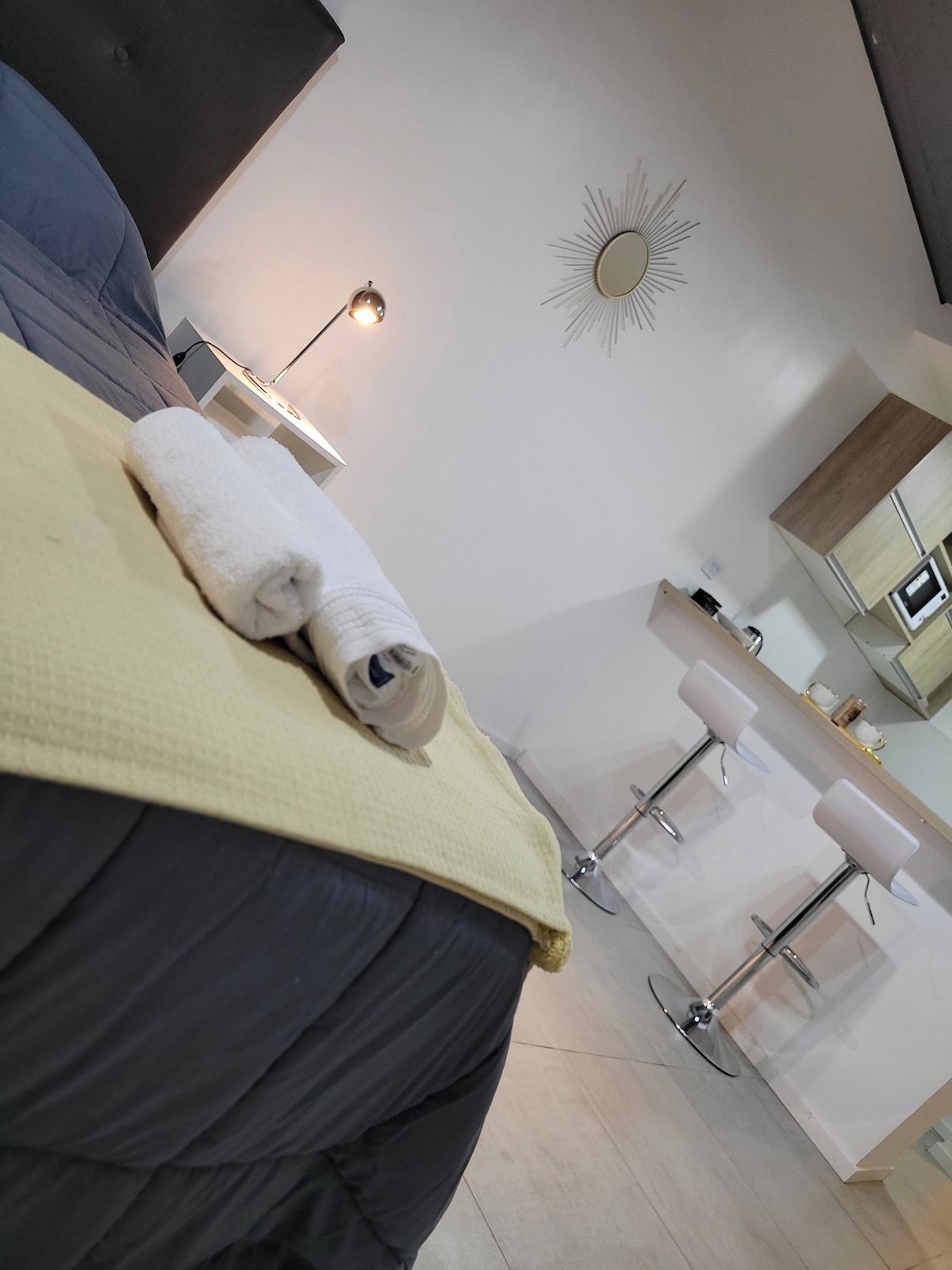 A modern studio is showcased with a neatly made spacious bed, accented by a light yellow blanket. Clean white towels are rolled and placed on the bed. In the background, a well-lit kitchen area with bar stools and a decorative mirror adds to the inviting atmosphere.
