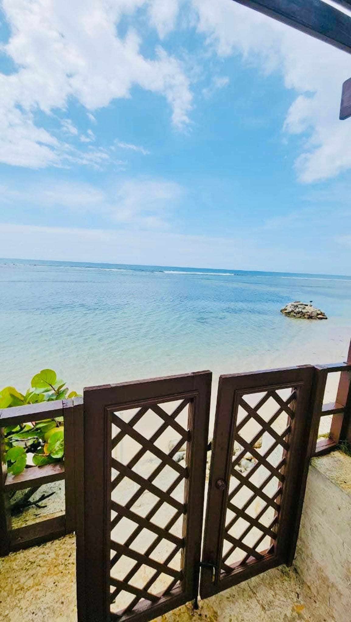 A pair of wooden gates opens to a serene beachfront scene, showcasing calm waters that reflect the blue sky. Lush greenery frames the entrance, leading to sandy shores and gentle waves in the distance.