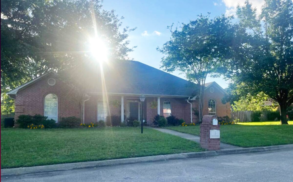 A brick house is situated in a serene neighborhood, framed by lush green grass and well-maintained shrubs. Sunlight filters through leafy trees, enhancing the inviting outdoor space. A pathway leads to the front door, and a mailbox stands near the street.