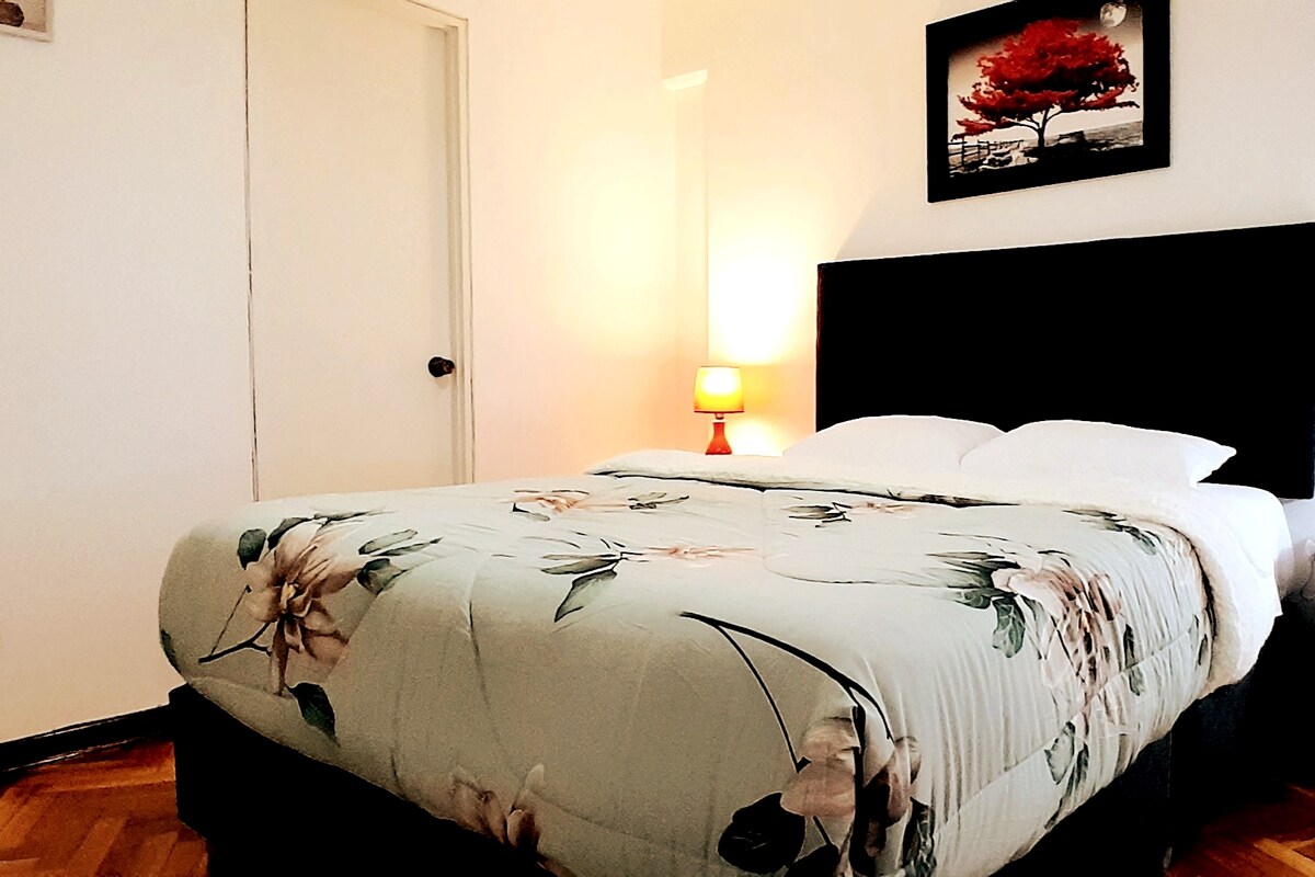 A spacious bedroom features a large bed adorned with a floral-patterned blanket and white pillows. A bed lamp emits soft light beside the bed, while a framed artwork depicting a tree is displayed on the wall.