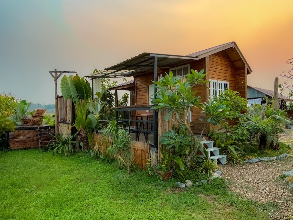 A charming wooden house is nestled within lush greenery, featuring large windows and a sloped roof. A small set of steps leads to the entrance, while a cultivated garden with various plants surrounds the property, enhancing its serene and natural setting.
