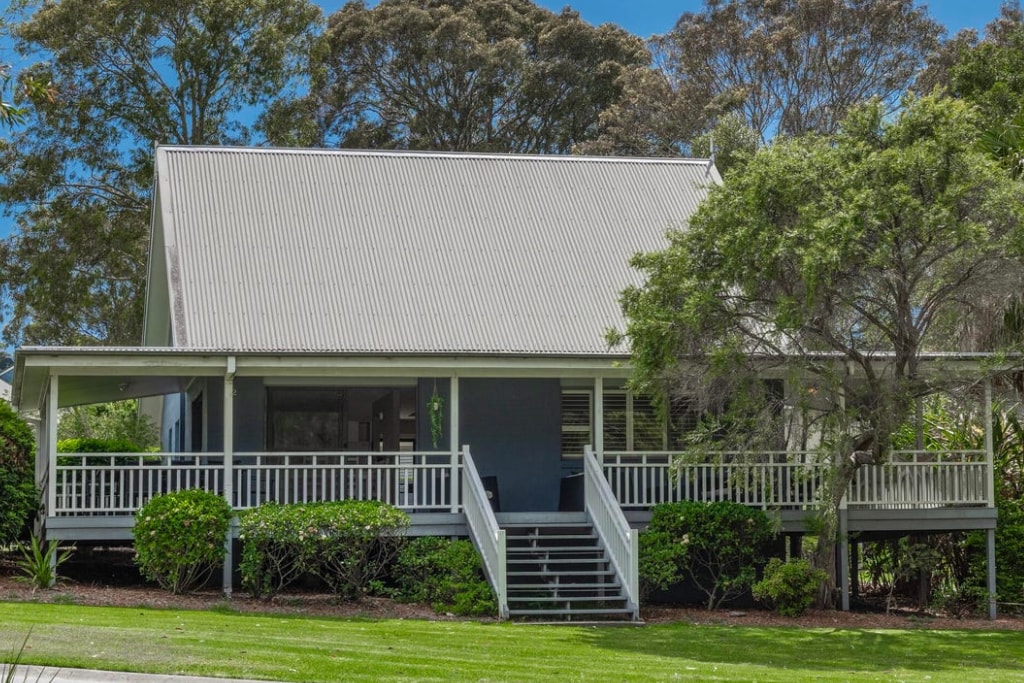 The exterior of a three-bedroom holiday house is visible, featuring a sloping metal roof and a spacious front porch surrounded by well-maintained greenery. A pathway leads to the entrance, and large windows provide a glimpse of the welcoming interior. Tall trees frame the property.