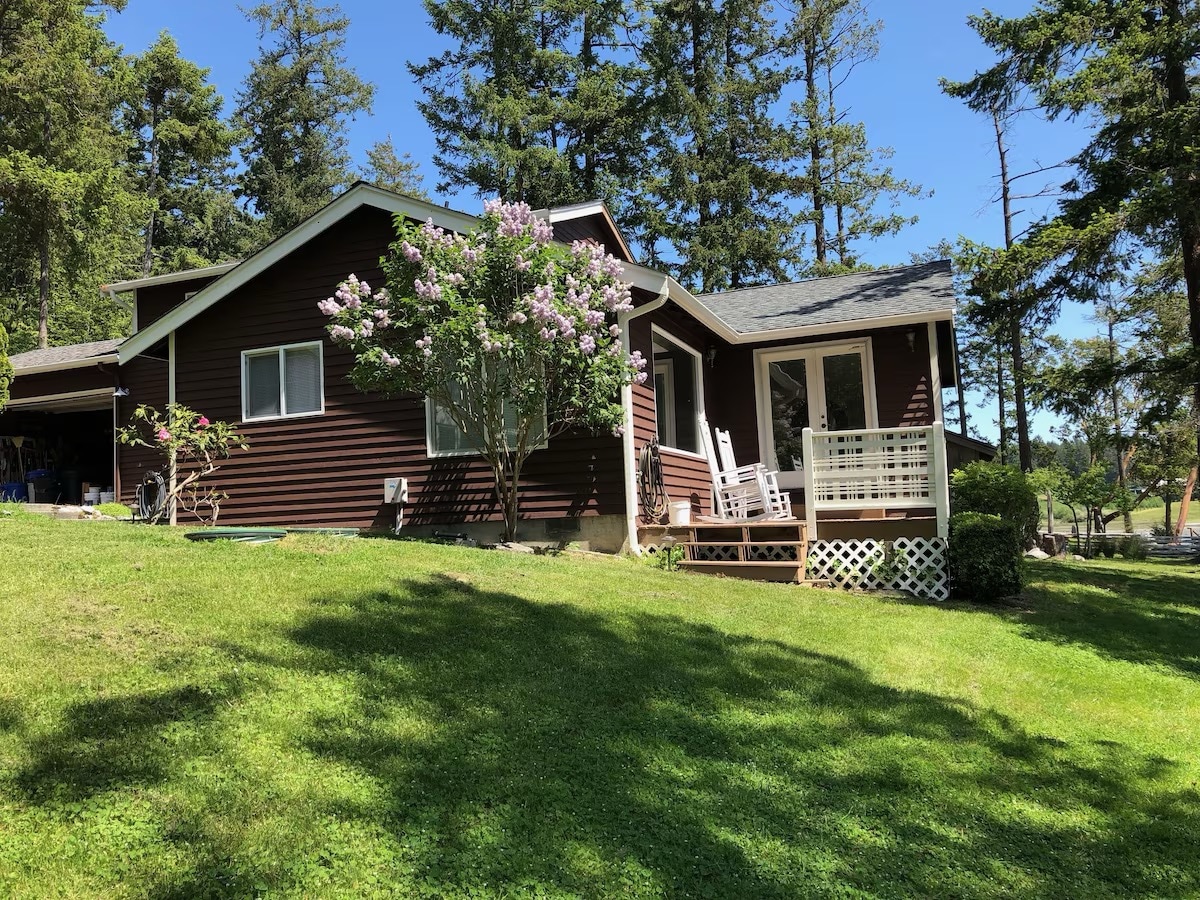 A brown wooden house is set on a grassy slope, surrounded by tall trees. A flowering bush adds color to the exterior, while a deck is visible, featuring white railing and rocking chairs. Sunlight casts gentle shadows across the well-kept lawn.