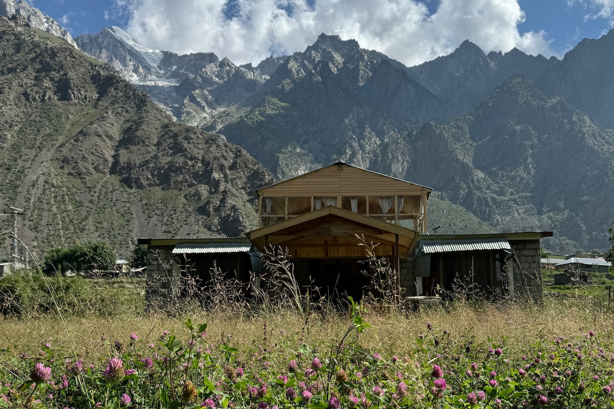 A two-story house is positioned amidst grassy grounds adorned with blooming wildflowers. Towering mountains serve as a dramatic backdrop, with peaks touched by snow under a partly cloudy sky.