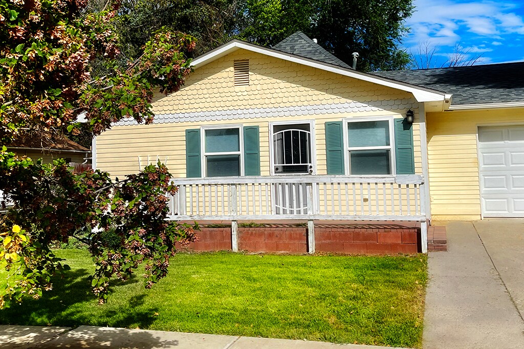 A yellow house featuring a welcoming front porch with a white railing and green shutters. A neatly maintained lawn is visible in front, complemented by a leafy tree. The garage door is located to the right of the entrance.
