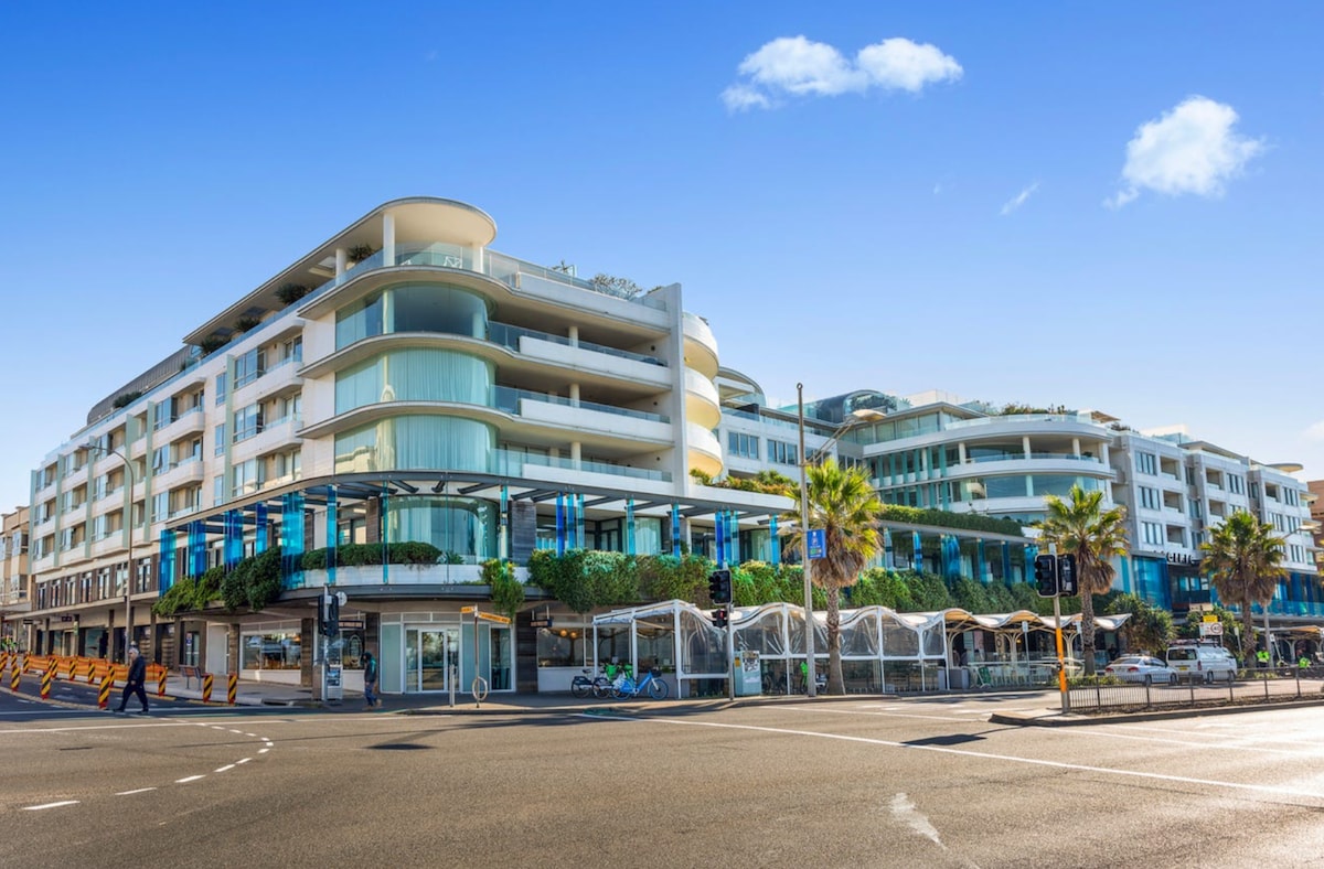A contemporary multi-story building is displayed, featuring a blend of glass and white exterior elements. Lush greenery and palm trees surround the ground level, while clear blue skies hover above, highlighting the inviting atmosphere of the Bondi Beach area.