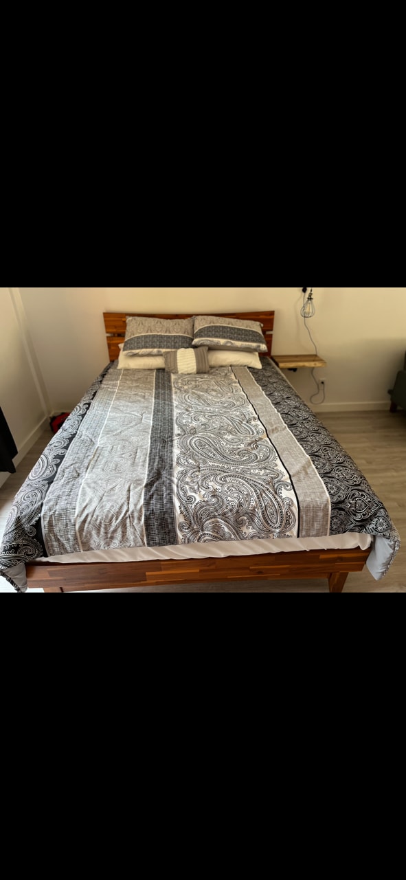 A spacious bed features an intricate patterned coverlet in neutral tones. Two neatly arranged pillows are positioned at the headboard, with a small bedside table visible adjacent to the bed.