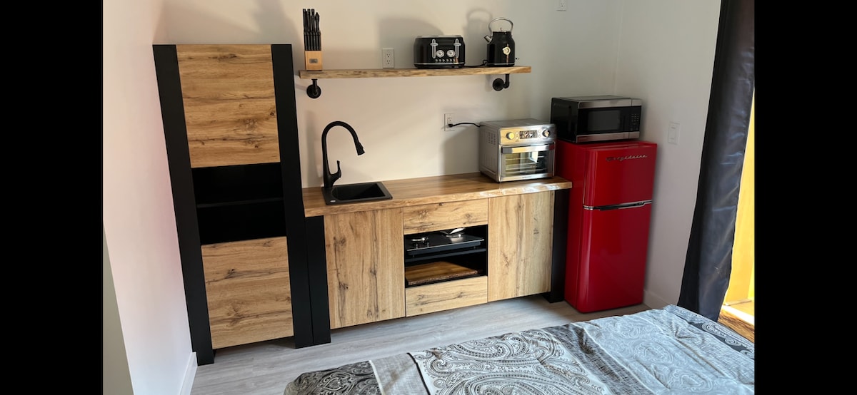 A compact kitchen area is presented, featuring a wooden countertop with a black sink and a small appliance area. A red refrigerator stands alongside a microwave and a toaster oven. Shelves store kitchenware, contributing to the organized and functional space.