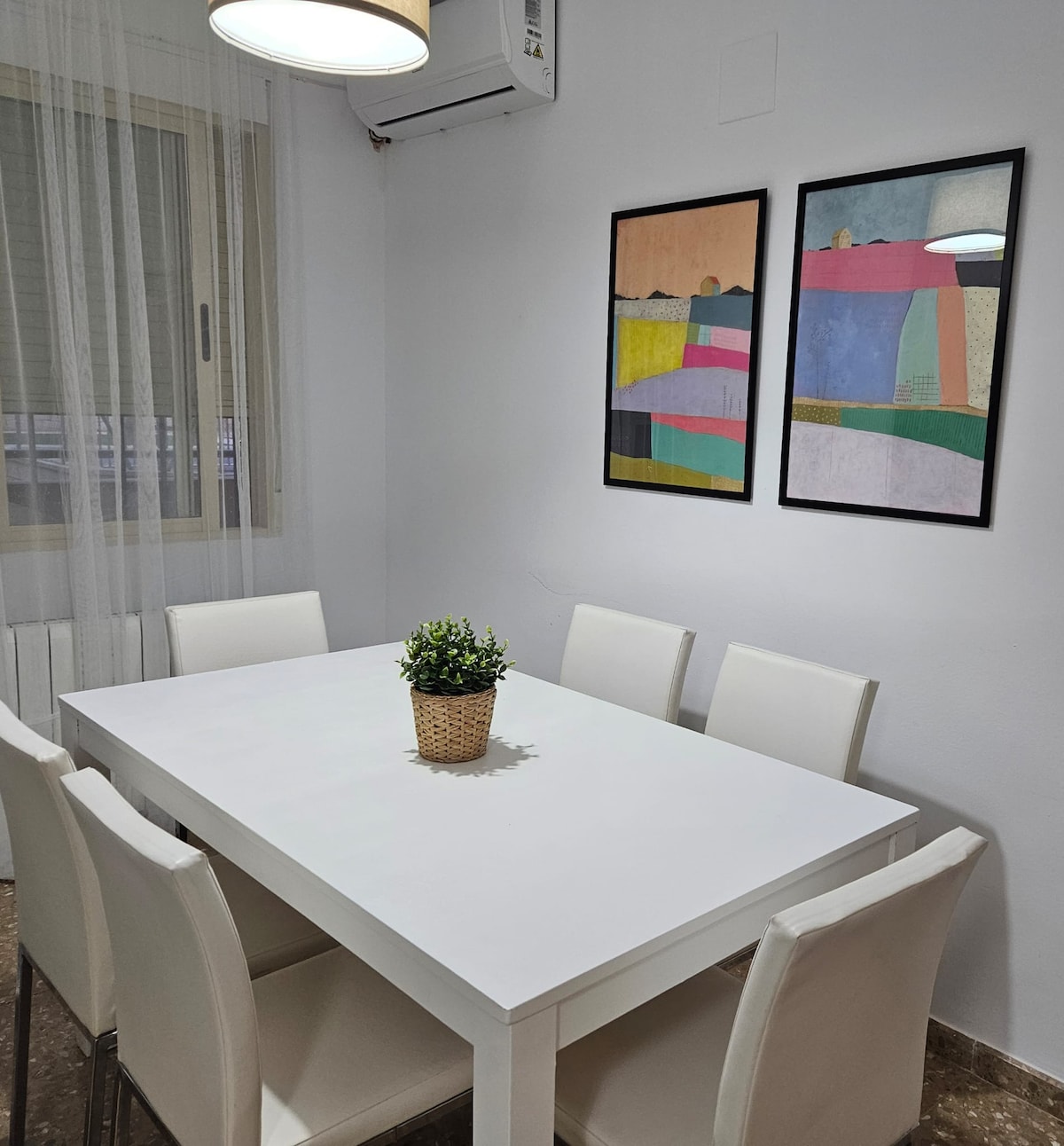 A bright dining area is equipped with a white table surrounded by six light-colored chairs. Two framed art pieces adorn the wall, adding a touch of color to the space. Natural light enters through a window with a sheer curtain.