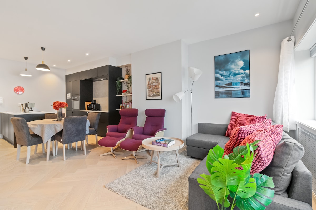 An open-concept living area features a cozy gray sofa and two purple reclinable armchairs, complemented by a small wooden table. A dining table is visible in the background, surrounded by chairs. Decorative plants and a colorful wall art contribute to a welcoming environment.