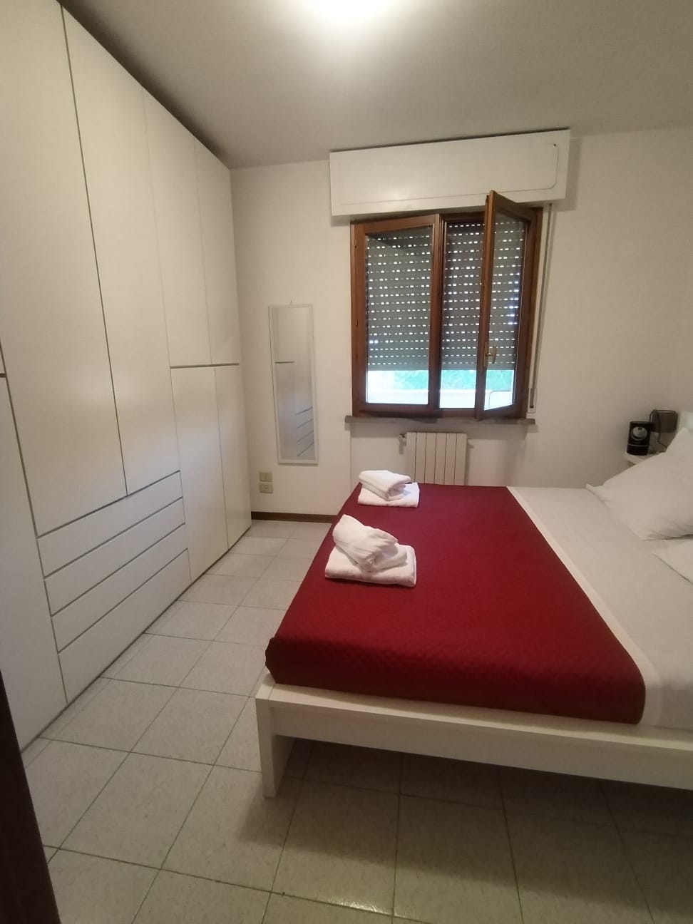 A bedroom features a spacious bed with a red blanket, complemented by neatly arranged white towels. A large wardrobe provides ample storage, while natural light filters through the partially open window dressed with wooden blinds. A mirror is positioned against the wall, enhancing the room's functionality.