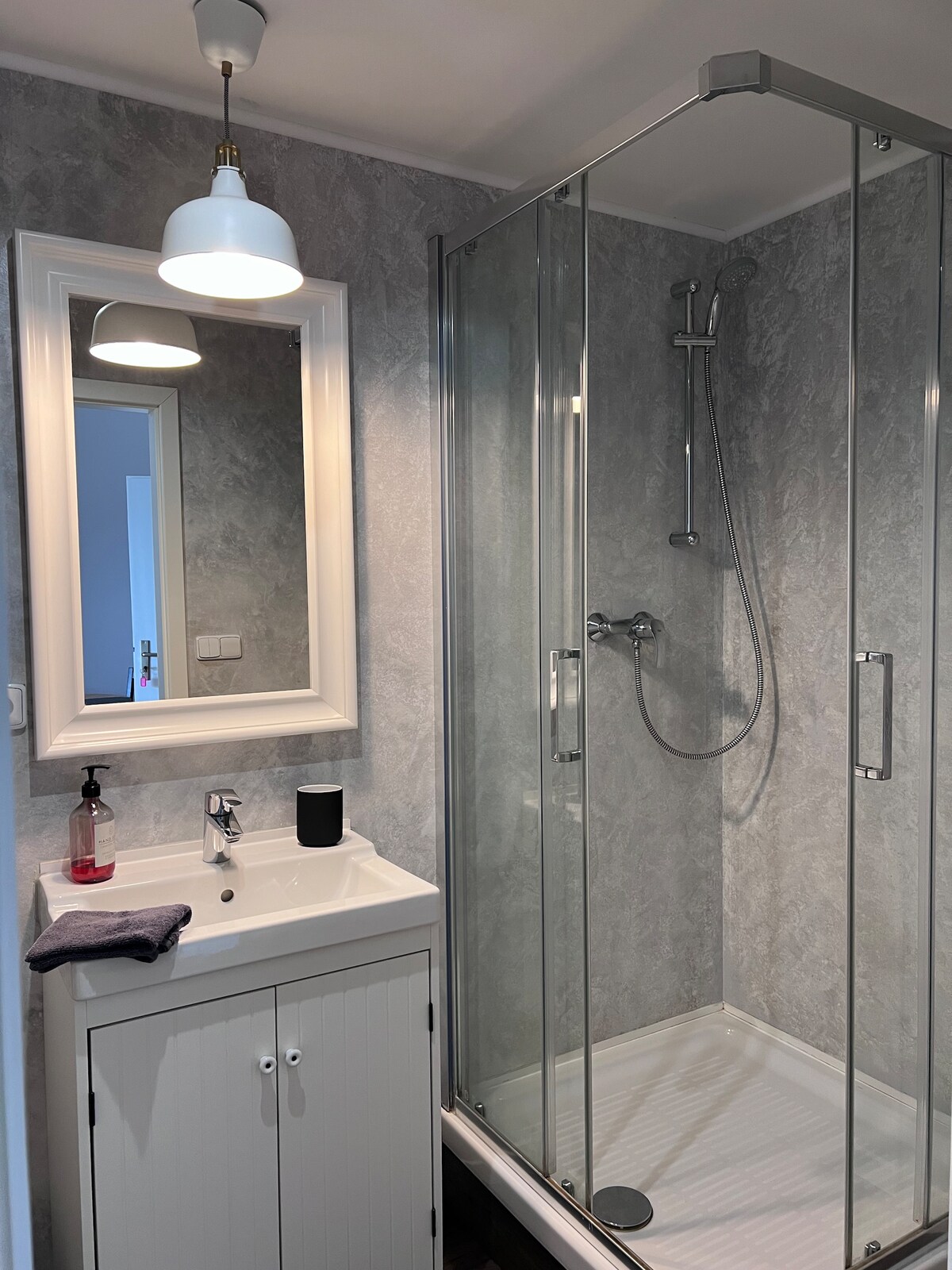 A well-appointed bathroom features a glass shower enclosure, showcasing modern fixtures. A white vanity with storage cabinets is positioned beneath a framed mirror, reflecting a bright light fixture overhead. Neutral grey walls provide a subtle backdrop, creating a functional and clean space.