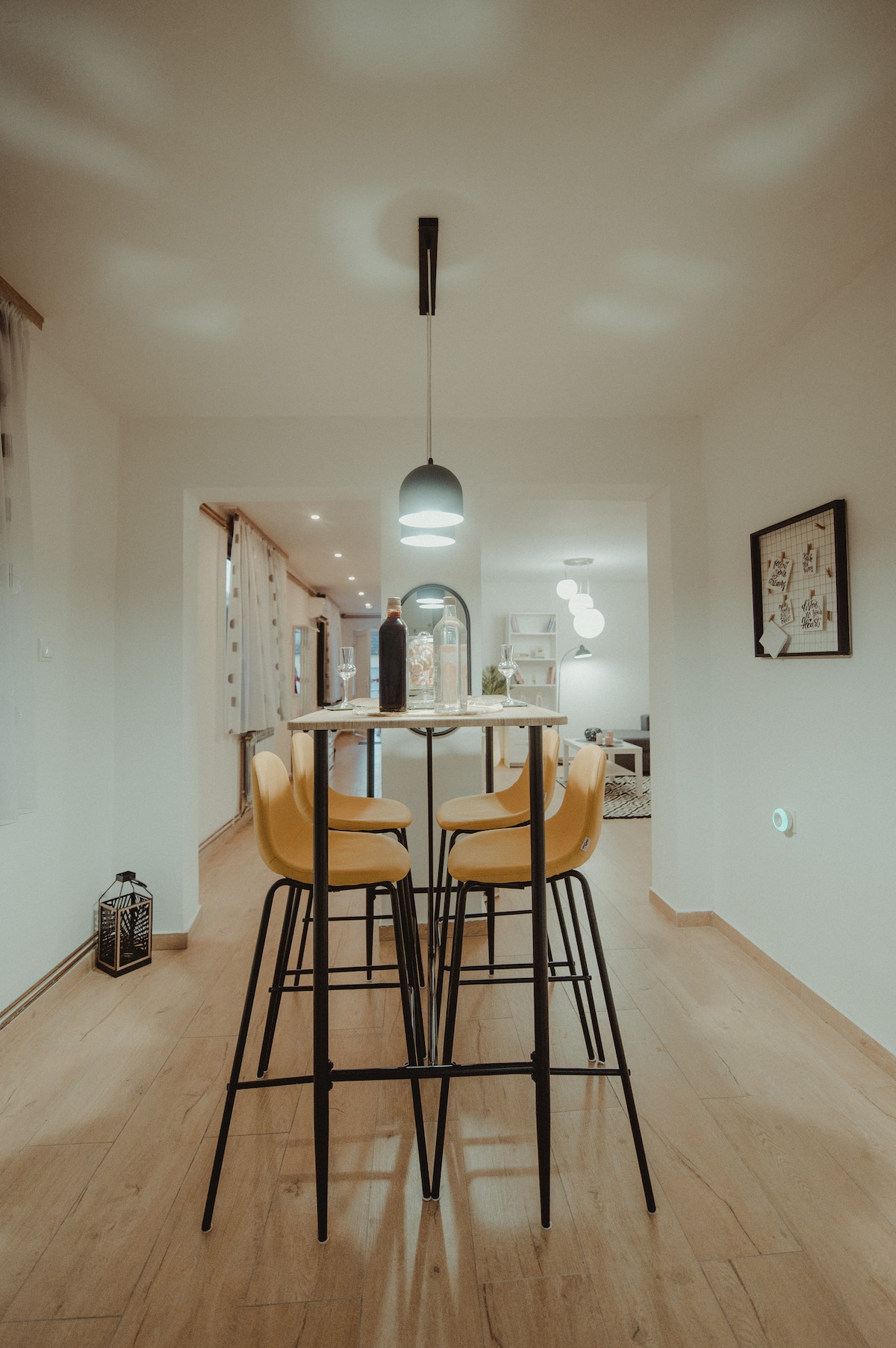 A dining area features a sleek table surrounded by yellow bar stools. A pendant light hangs above, casting soft shadows on the walls. The background reveals a glimpse of the living space, creating an open and inviting atmosphere.