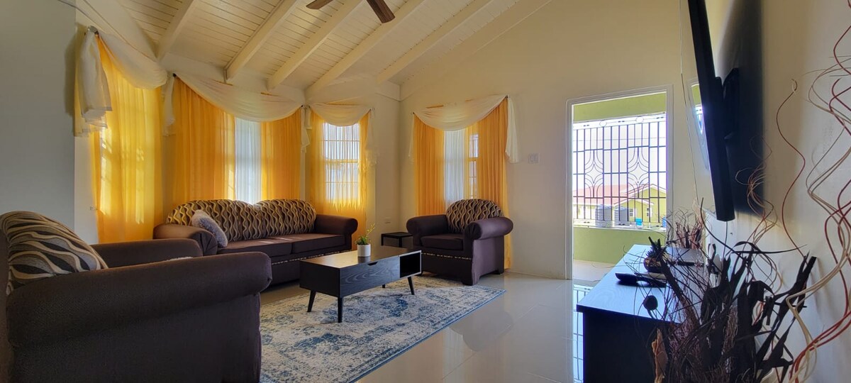 A bright living area features large windows dressed with flowing yellow curtains. Two couches and a lounge chair surround a central coffee table atop a patterned area rug. A flat-screen television is mounted on a light-colored wall, enhancing the open and airy feel of the space.