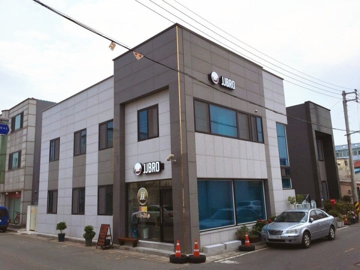 A modern two-story building with a sleek grey and black exterior features large windows that allow ample natural light. The entrance is marked by a circular sign displaying the name 'JJBRO,' while potted plants line the entrance, enhancing the welcoming appearance.