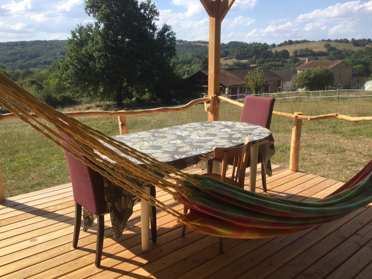 A covered terrace features a wooden table surrounded by four chairs, complemented by a colorful hammock draping across the space. Expansive views of rolling hills and greenery are visible in the background, creating an outdoor setting that promotes relaxation.