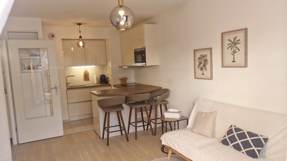 An open living area features a contemporary kitchen with sleek cabinets and modern appliances. A dining table with four stools is situated beside the kitchen. A light-colored sofa is placed in the corner, decorated with a patterned cushion. Framed botanical prints adorn the wall.