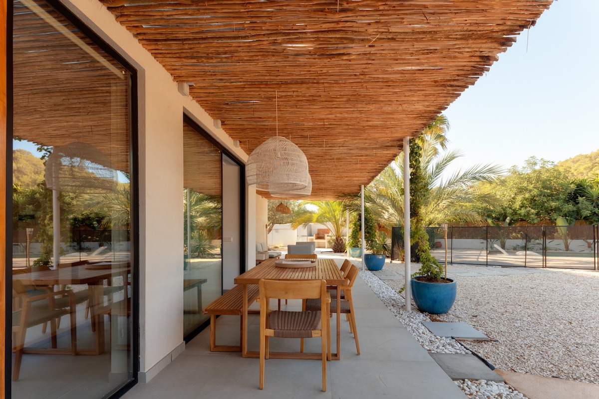 An outdoor dining area is featured under a natural wood awning, providing shade. A large wooden table is surrounded by comfortable seating, creating an inviting space. Lush greenery is visible in the background, enhancing the serene atmosphere. Potted plants add color to the setting.