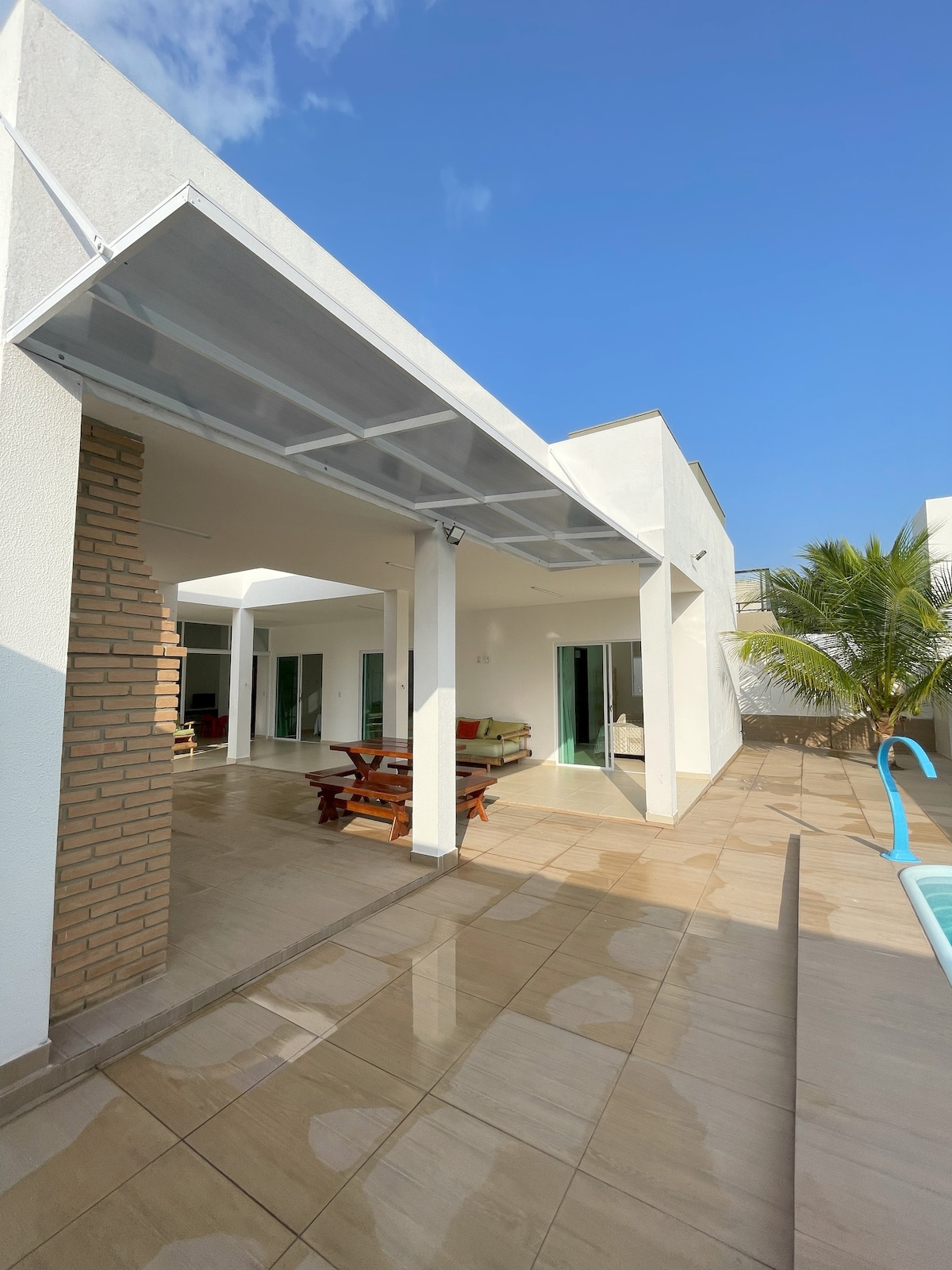 An outdoor area is depicted, featuring a spacious patio with tiled flooring and a dining table made of dark wood. Sunlight reflects off the surface, enhancing a bright atmosphere. Tropical plants are present, and a swimming pool can be seen in the corner, complementing the modern white architecture.