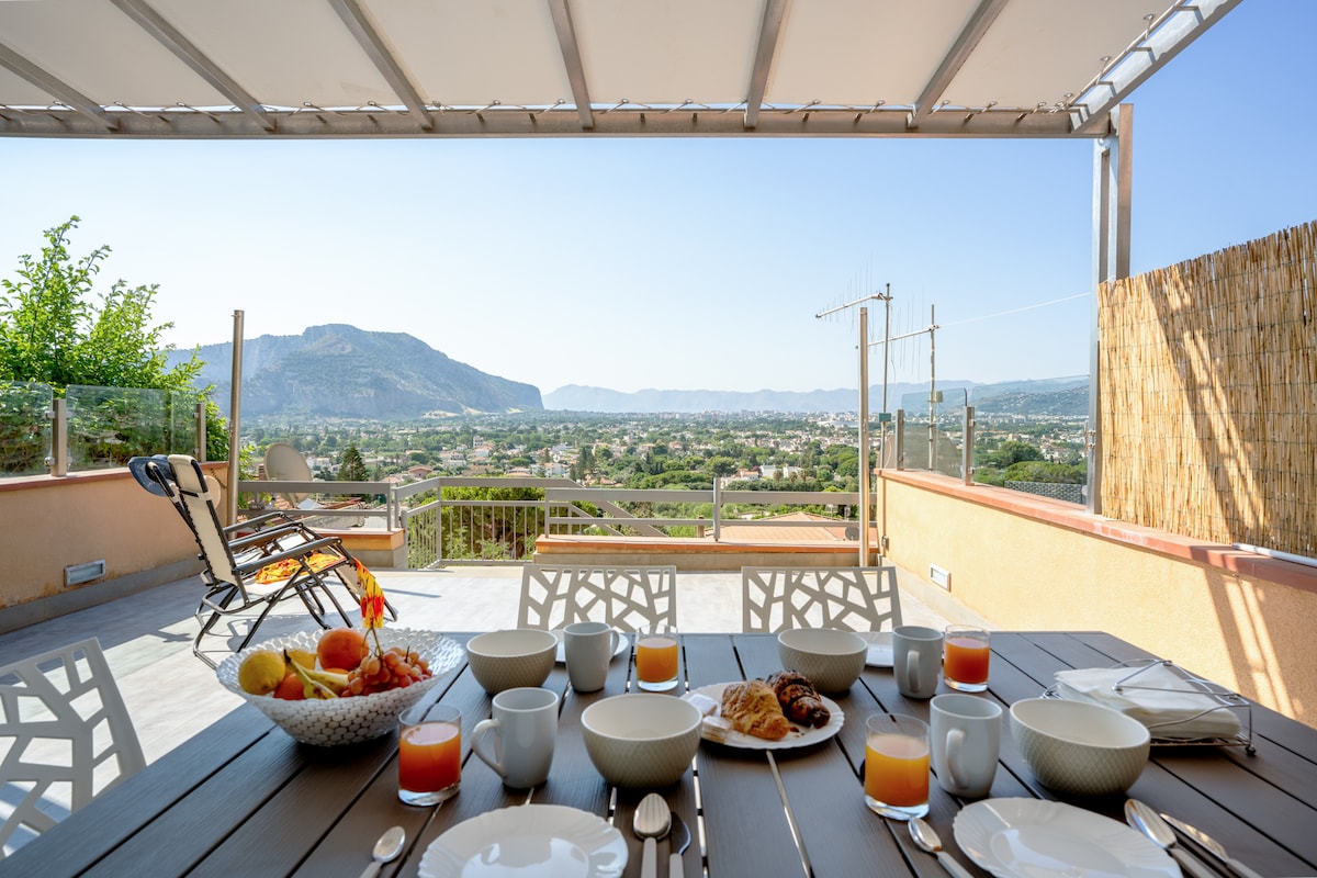 An outdoor terrace is set up for dining, featuring a large table with plates, bowls, and glasses arranged for breakfast. A view of the surrounding landscape and mountains can be seen in the background, complemented by a comfortable lounge chair nearby.