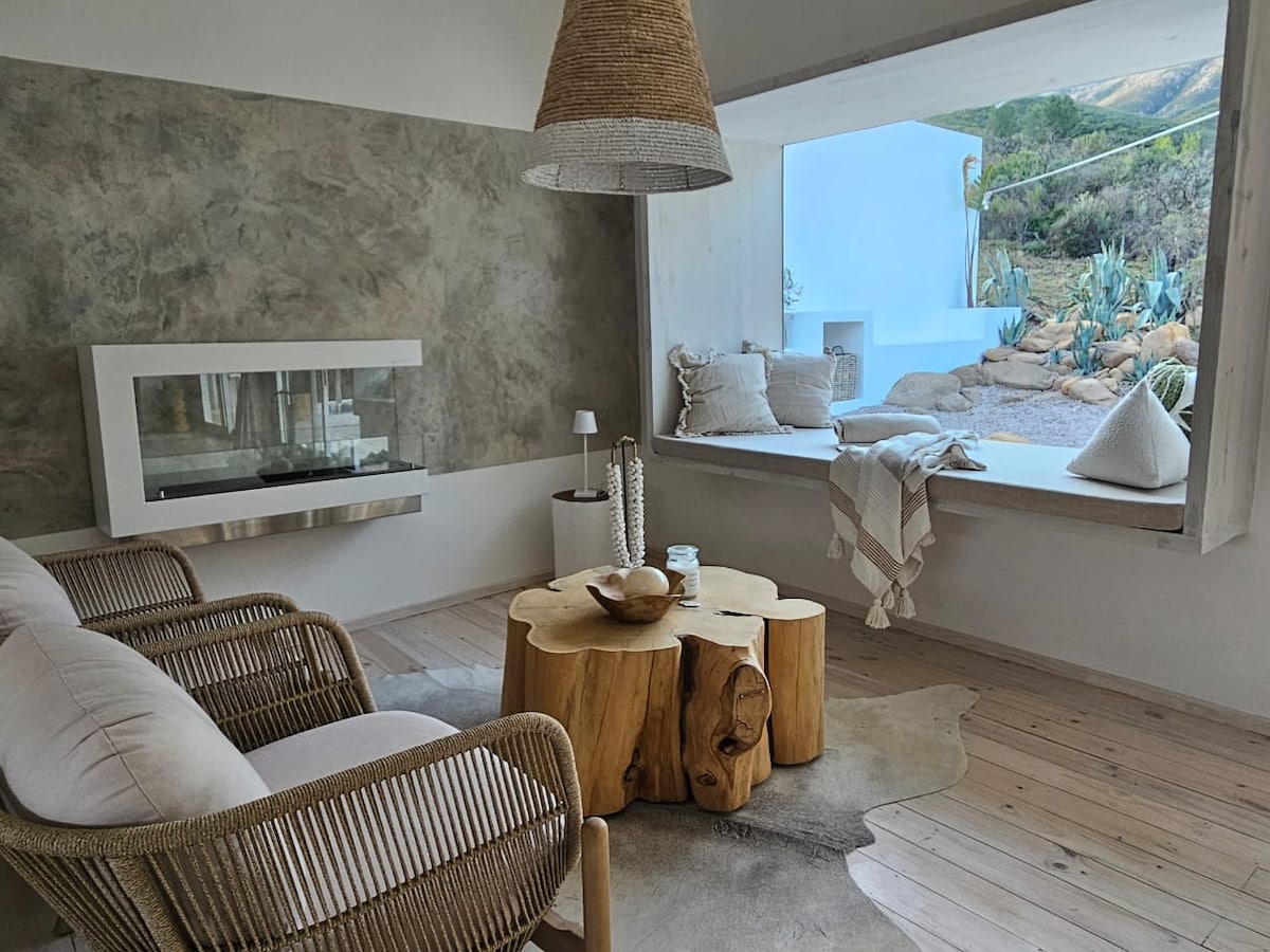 A cozy seating area features two comfortable armchairs made of natural materials, positioned beside a handcrafted wooden coffee table. A modern fireplace is integrated into the wall, while a large window provides a scenic view and seating with decorative pillows.