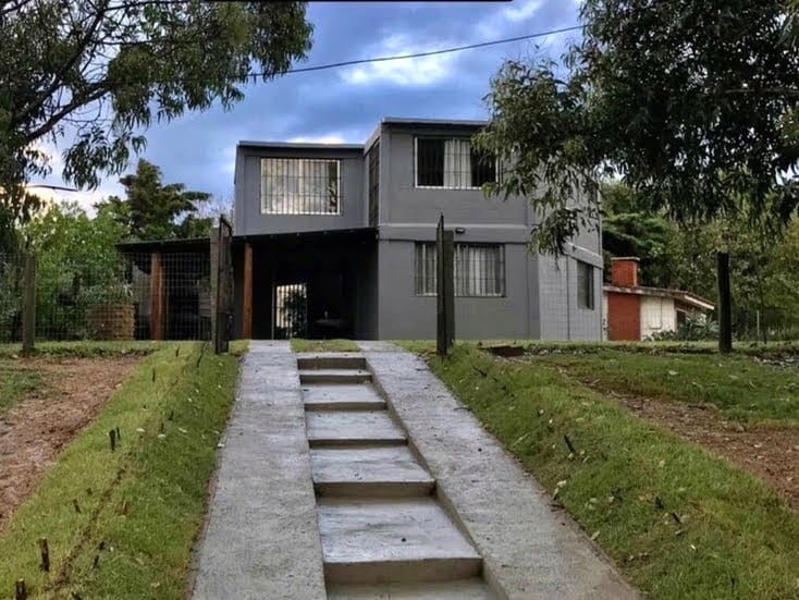 The exterior of the house is showcased, featuring a two-story structure set within a green environment. A concrete path leads to the entrance, framed by grass and trees. Large windows provide a view into the interior from the front.