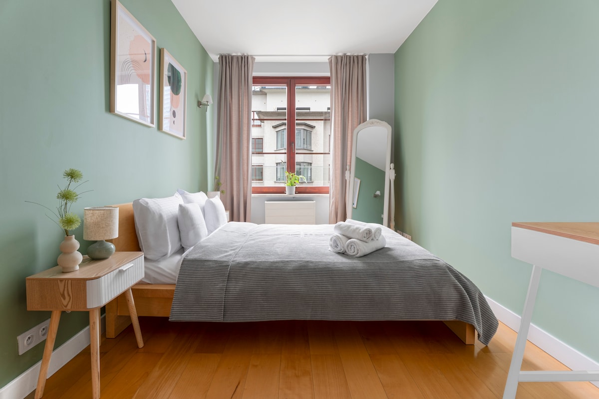 The bedroom features soothing green walls and a spacious bed with crisp white linens and neatly arranged towels. Natural light enters through a large window adorned with light curtains. A simple wooden nightstand complements the bed, while a mirror stands gracefully across the room.