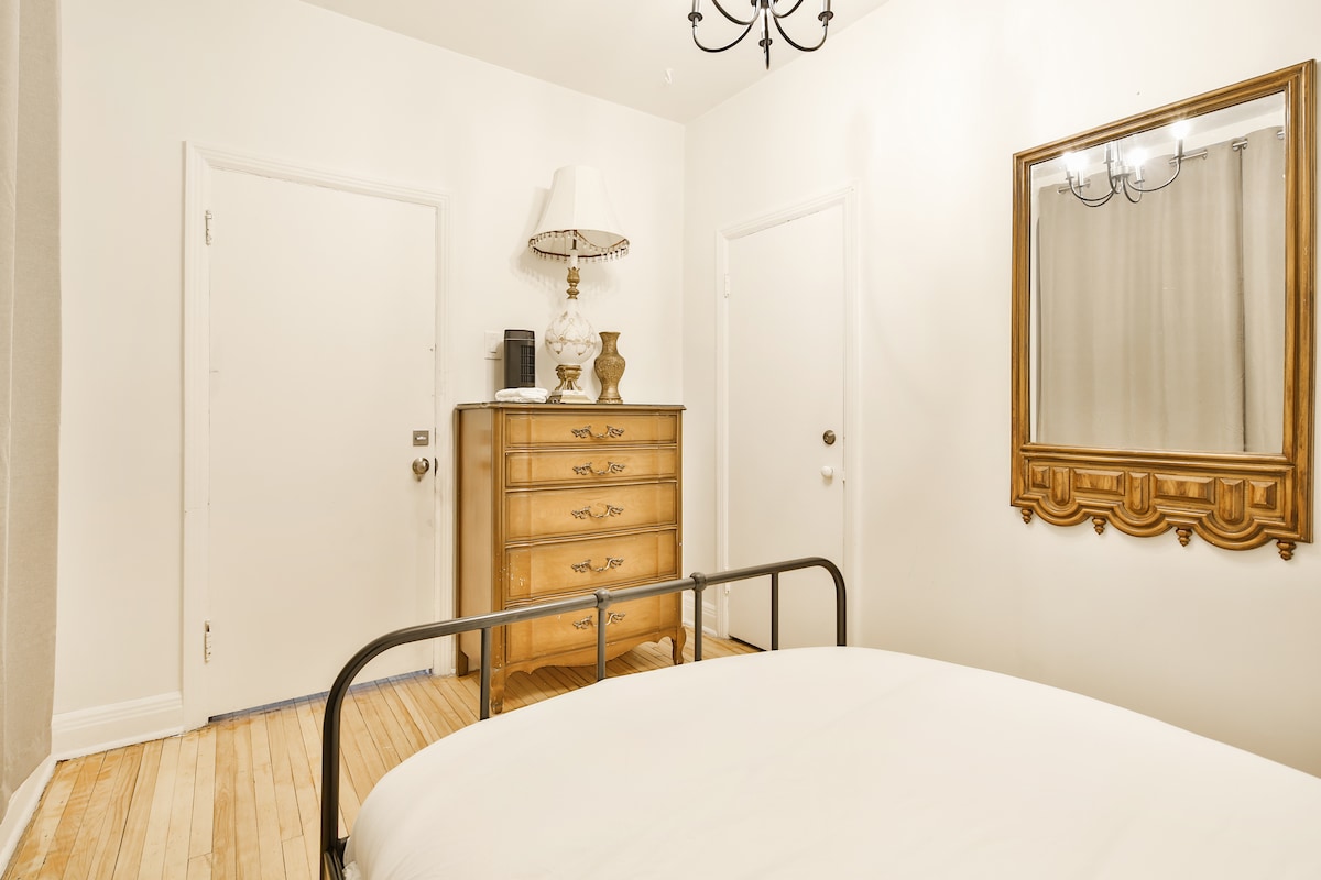 A private bedroom features a spacious bed with crisp white linens. A wooden dresser, adorned with decorative items, stands against the wall. A vintage mirror reflects light from a nearby lamp, while simple doors lead to the hallway and bathroom.