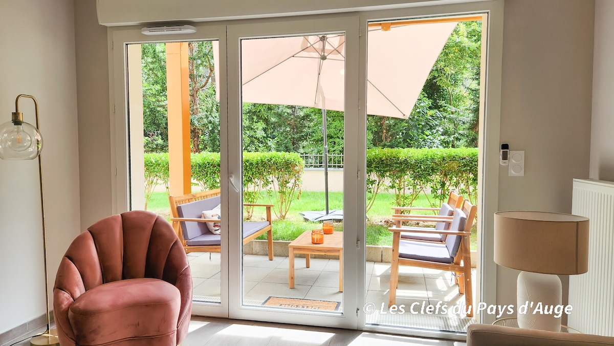 A spacious living area features large glass doors that open to a small garden. The outdoor space is furnished with two wooden chairs and a coffee table, shaded by an umbrella. Lush greenery surrounds the area, enhancing the inviting atmosphere.