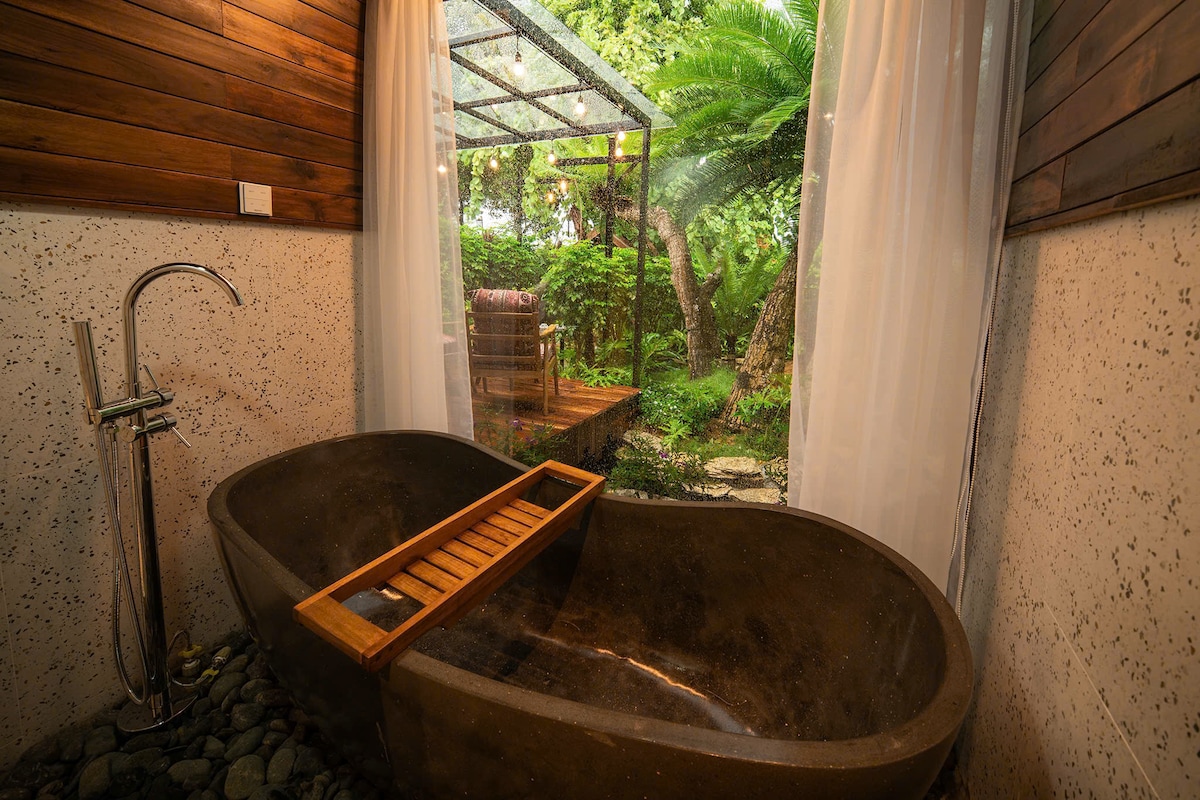 A freestanding bathtub is positioned next to a large window, providing a view of the lush greenery outside. Natural light filters through sheer curtains, highlighting the stone-like texture of the tub and the wooden slat tray resting on the edge.