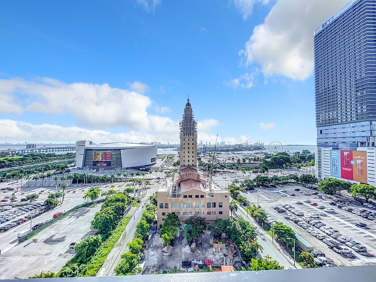 A panoramic view captures the iconic Freedom Tower surrounded by green landscaping and urban spaces. Nearby, modern buildings and open parking lots are visible, under a clear blue sky with a few clouds. The proximity to major attractions adds to the vibrant city atmosphere.