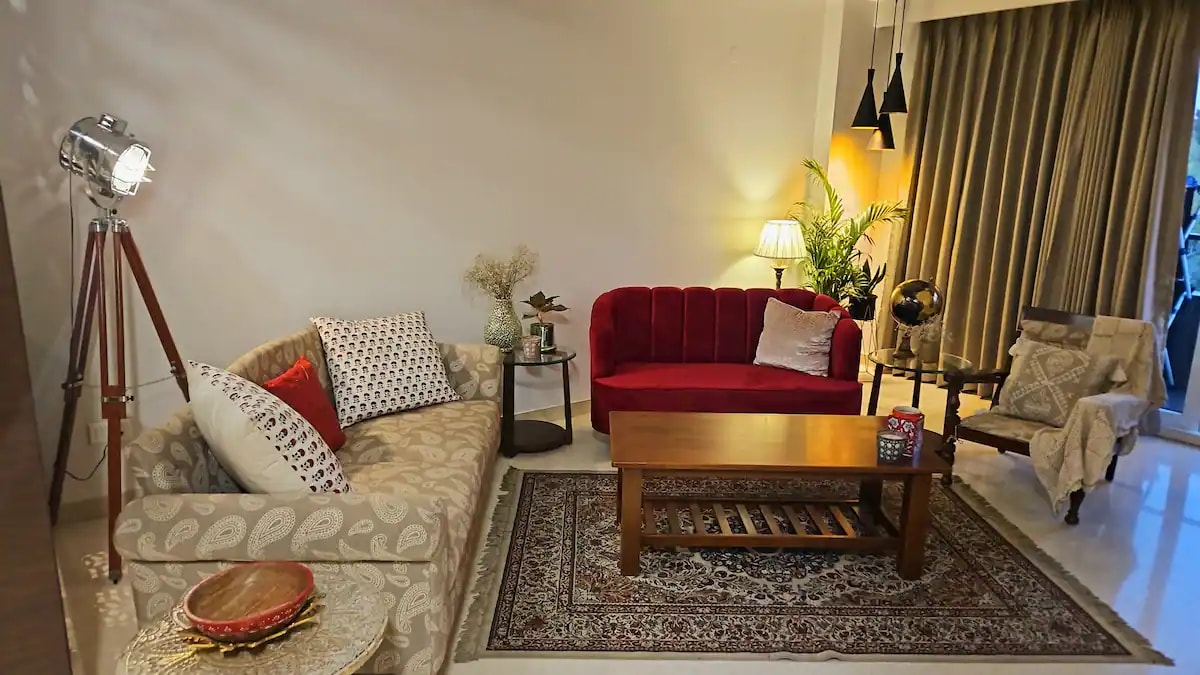 A living area features a light-colored sofa with decorative cushions and a vibrant red loveseat. A wooden coffee table sits centrally on a patterned rug, and a plant adds a natural touch. Soft lighting is provided by a floor lamp and stylish hanging fixtures.