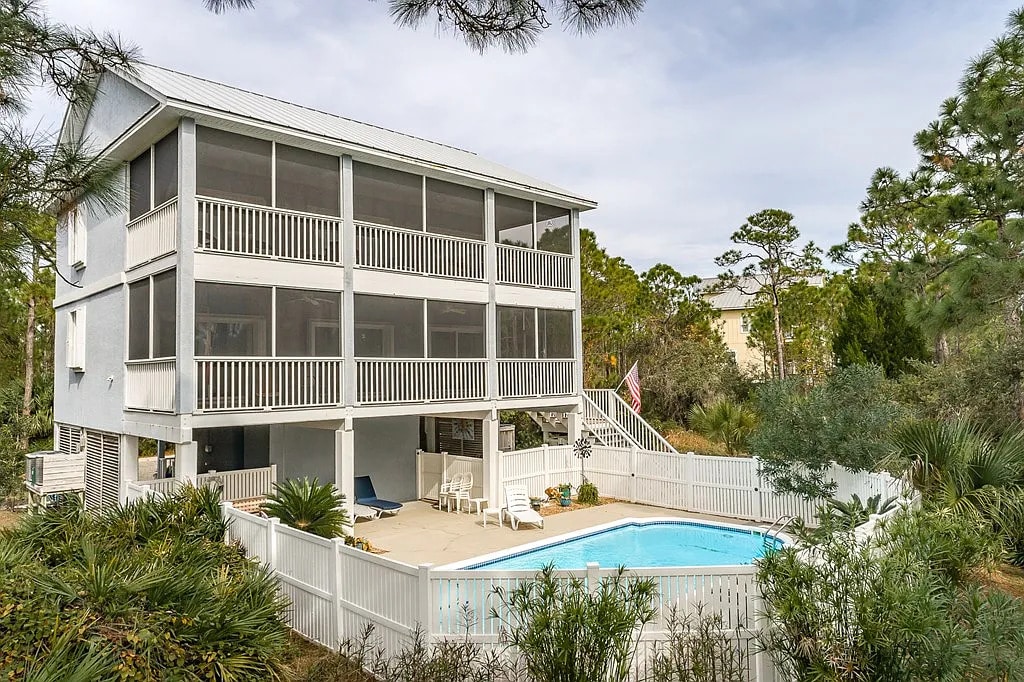 The exterior of the home is shown, featuring a multi-story structure with multiple screened porches and a spacious pool area. The private pool is surrounded by landscaping and a white fence, providing a serene environment. Tall trees and natural vegetation frame the scene.