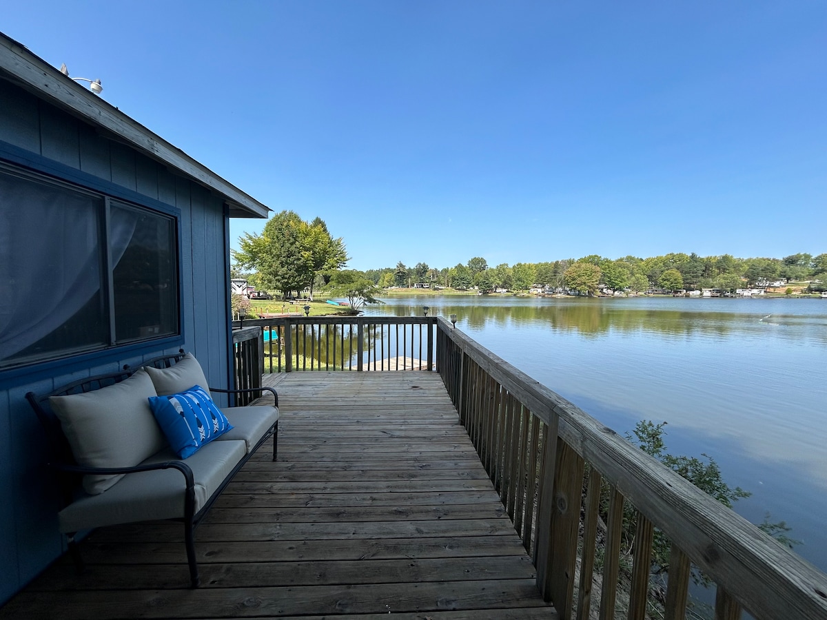 A spacious deck overlooks a serene lake, providing an inviting spot for relaxation. A comfortable seating area features a cushion-backed chair, with a decorative blue pillow. Lush green trees line the opposite shore, enhancing the tranquil view under clear skies.