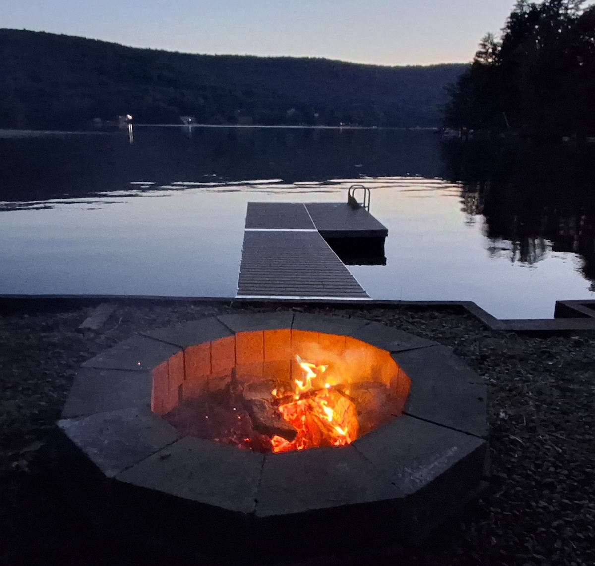 A stone firepit sits on the shoreline, with flames flickering gently. The calm lake reflects the twilight sky, while a dock extends into the water. Trees line the background, providing a serene setting for evening gatherings.