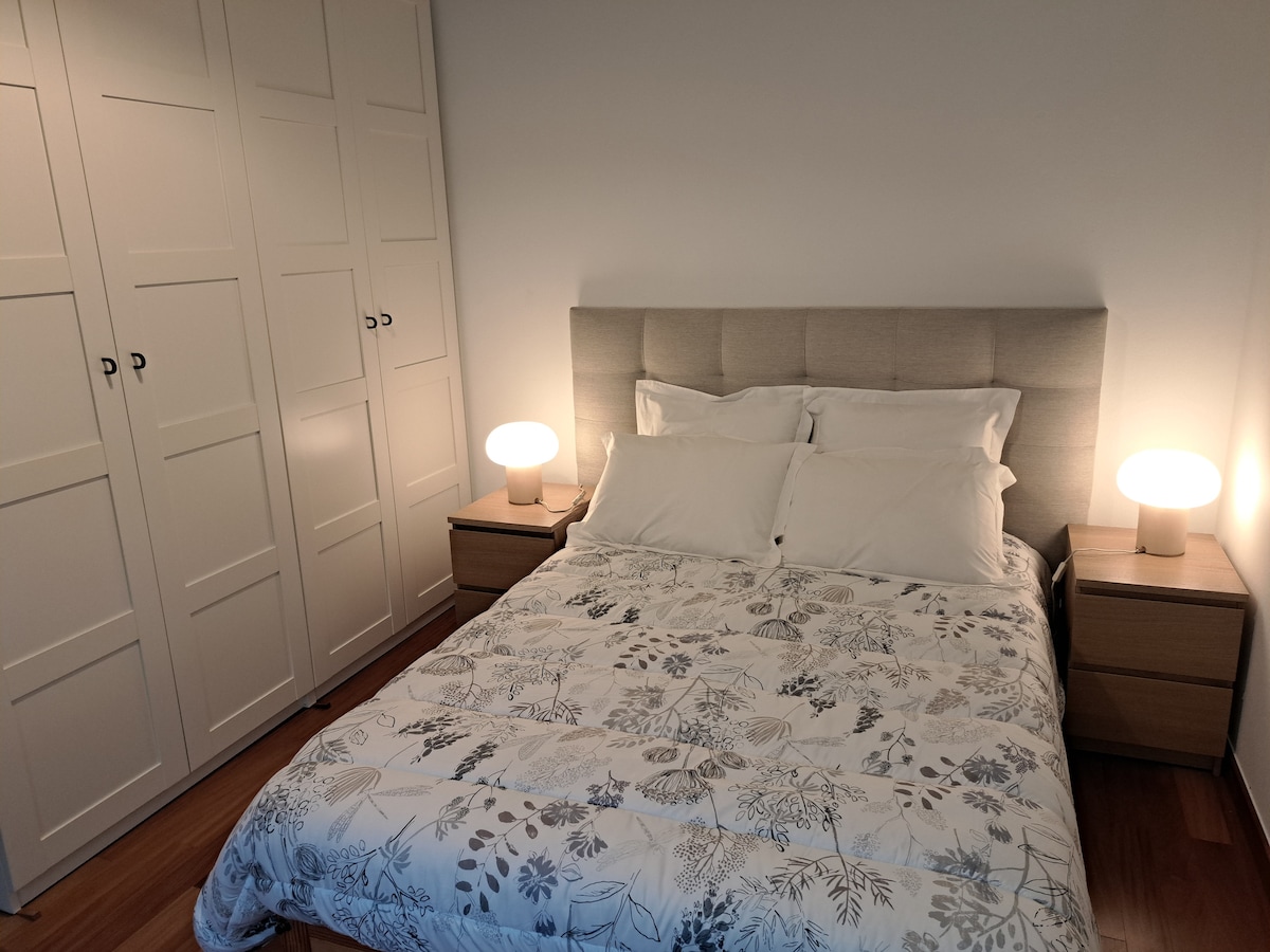 A cozy sleeping area is visible, featuring a spacious bed with a floral-patterned duvet and multiple pillows. Soft bedside lamps offer gentle illumination. A wardrobe with multiple panel doors is present in the background, enhancing the simplicity of the space.