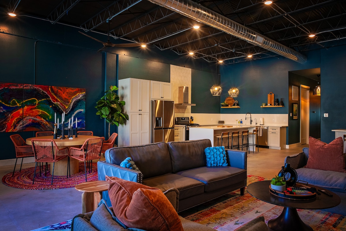 An open-concept living area features a modern kitchen with stainless steel appliances and a dining table surrounded by stylish chairs. A comfortable seating arrangement with colorful pillows adds to the inviting décor, complemented by a vibrant piece of wall art and warm lighting.