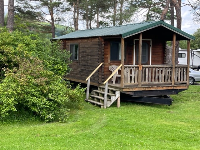 A cedar log cabin is surrounded by greenery, featuring a covered front porch with steps leading to the entrance. The rustic exterior blends with the natural landscape, while a white chair is positioned on the porch. Trees are visible in the background.