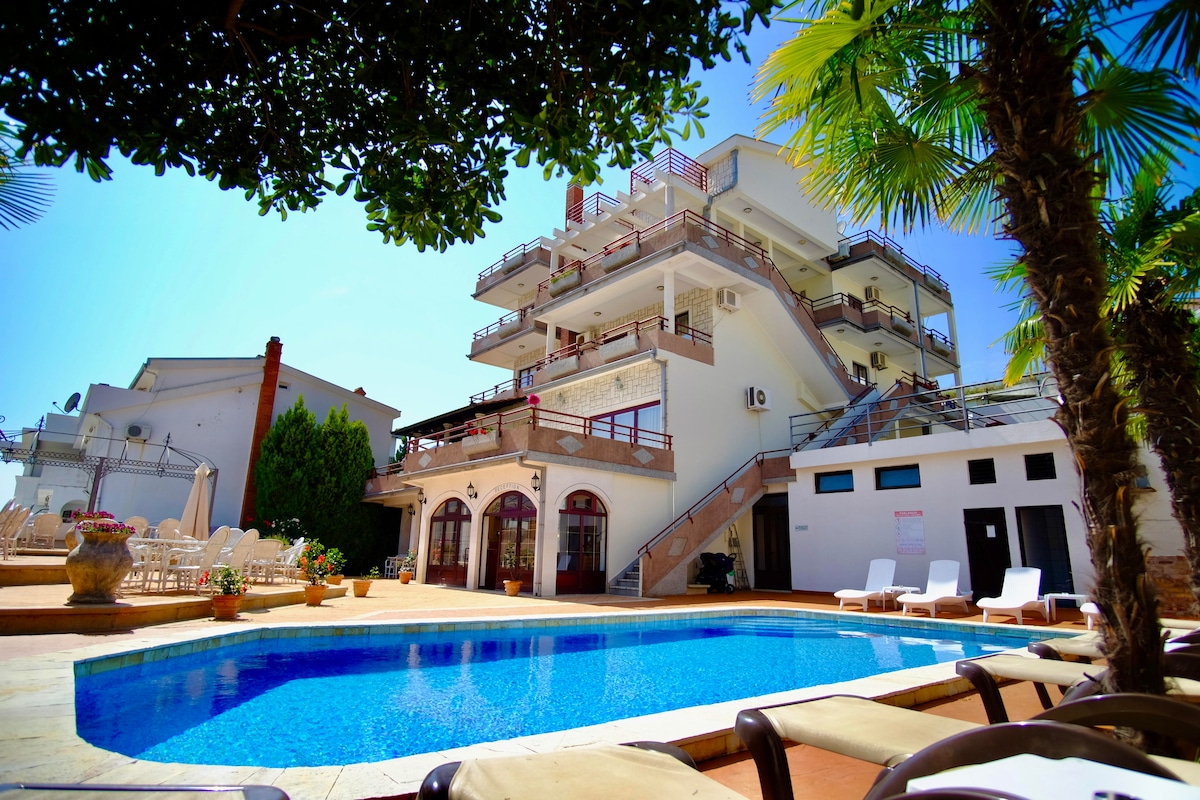 The image depicts a multi-story hotel building featuring numerous balconies surrounded by lush greenery. An inviting blue swimming pool is centered in the foreground, with sun loungers placed around its perimeter. Clear skies create a bright and open atmosphere.