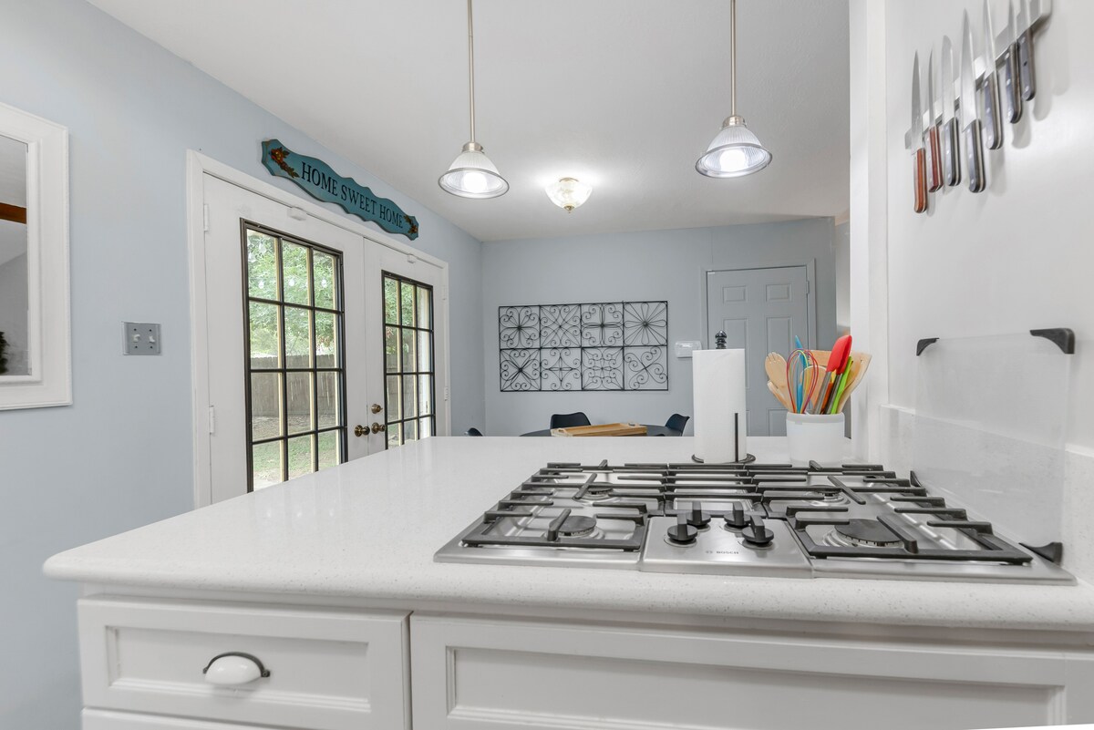 A bright and functional kitchen features a central island with a gas cooktop and modern white countertops. The wall is decorated with an array of knives, and the space is illuminated by pendant lighting. French doors lead to a view of the outdoors, enhancing the airy feel.