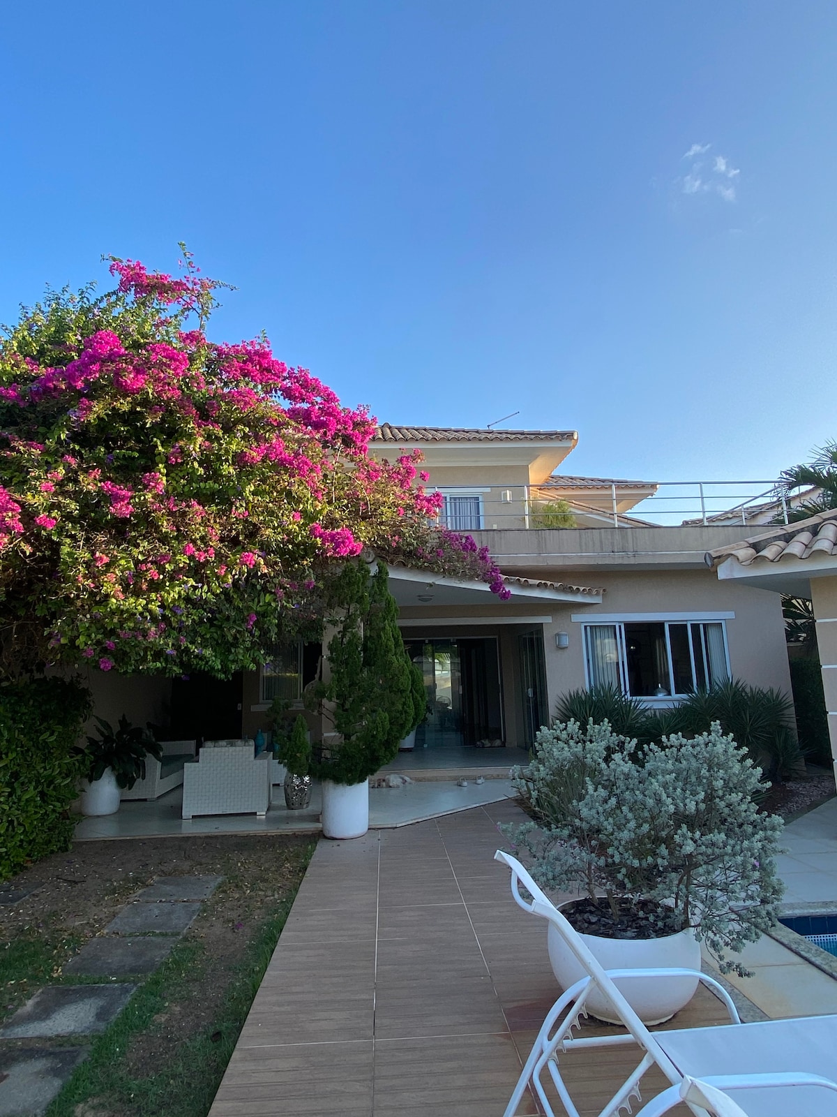 A spacious outdoor area is framed by vibrant, flowering plants and shrubs. The building features multiple stories with balconies and large glass doors leading indoors. Natural stone pathways connect the garden to the entrance, inviting exploration of the surrounding landscape.