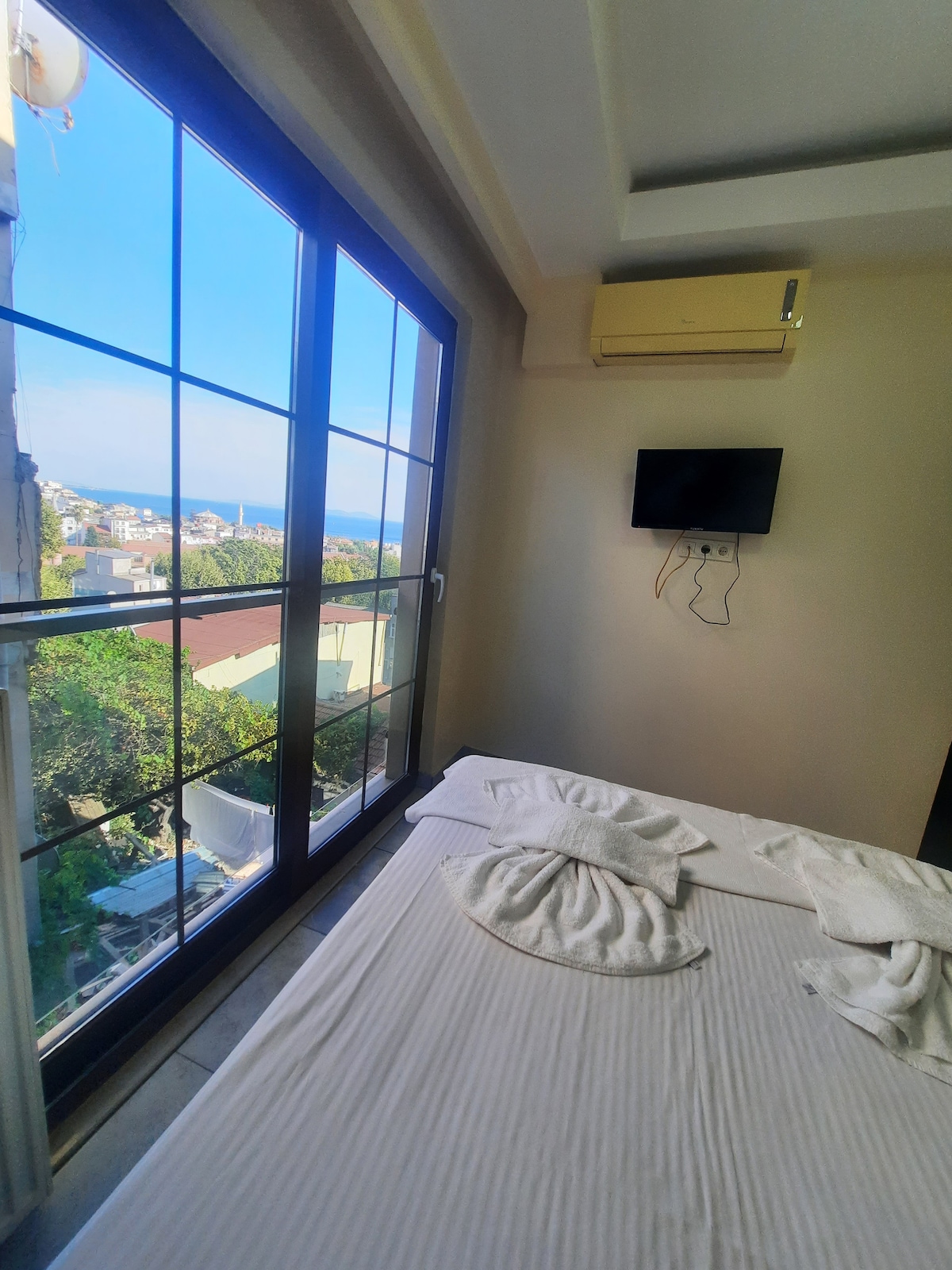 A cozy bedroom features a spacious bed with a light-colored bedspread. Large windows provide a view of the surrounding area and distant sea. An air conditioning unit is mounted above the bed, and a flat-screen TV is mounted on the wall opposite the window.