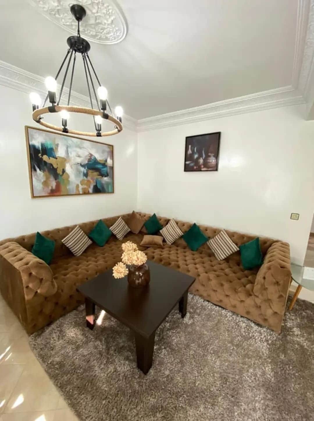 A modern living area features a large, plush tufted sectional sofa in warm tones, with decorative cushions in shades of green. A dark wooden coffee table sits centrally, adorned with a vase of dried flowers. Soft lighting from a stylish chandelier enhances the inviting space.