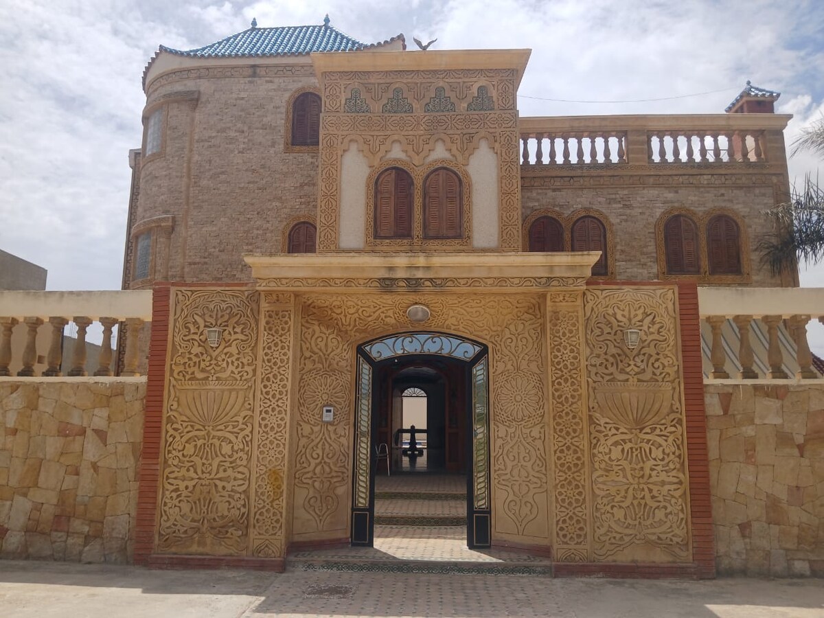 An ornate entrance features intricately carved stonework and traditional windows, leading into the villa. The doorway is framed by decorative arches, with a glimpse of the courtyard visible through the entrance. Surrounding walls showcase a blend of natural stone and stucco.
