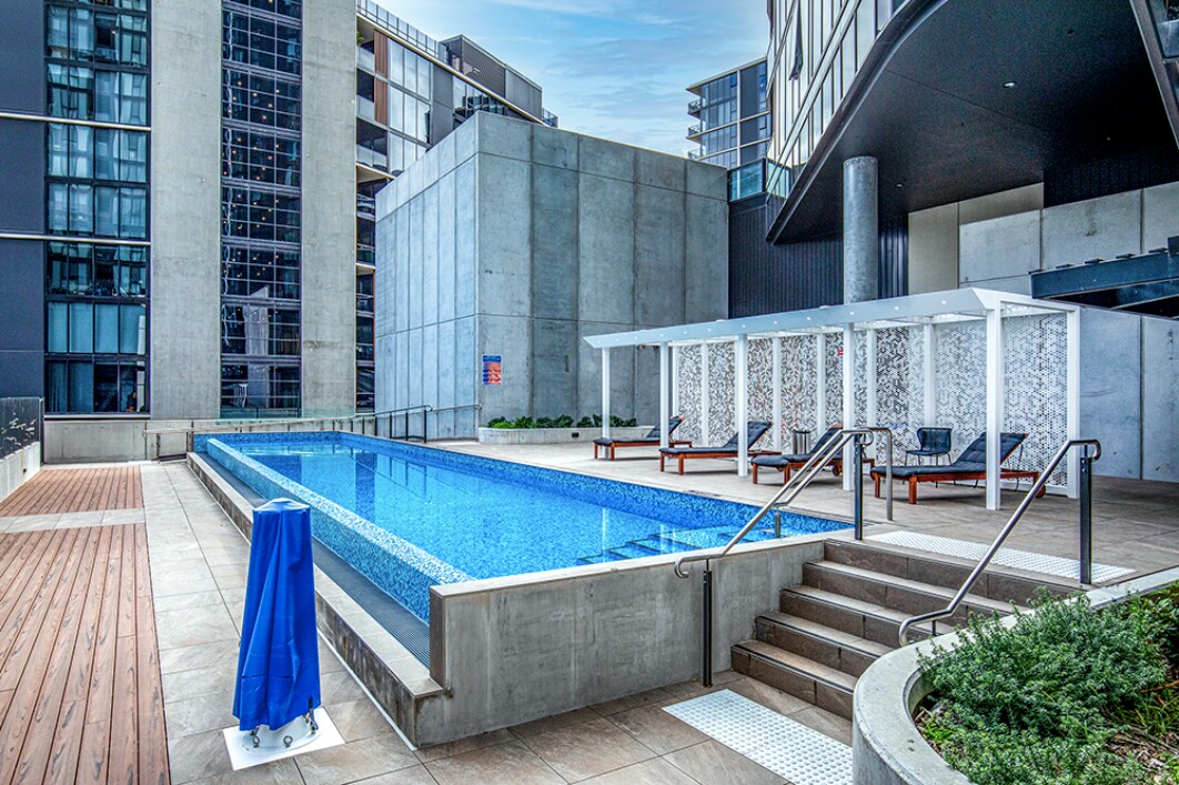 An outdoor pool area is displayed, featuring a clear blue water pool surrounded by modern architecture. Loungers are positioned adjacent to the pool, and a shaded cabana area is visible at one end, providing a relaxing space for guests.